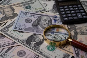 Aufnahme von zahlreichen Dollarscheinen, die auf einem Tisch liegen, zusammen mit einer Lupe und einem Taschenrechner – Symbolbild zum Geldanlageverhalten in Deutschland.