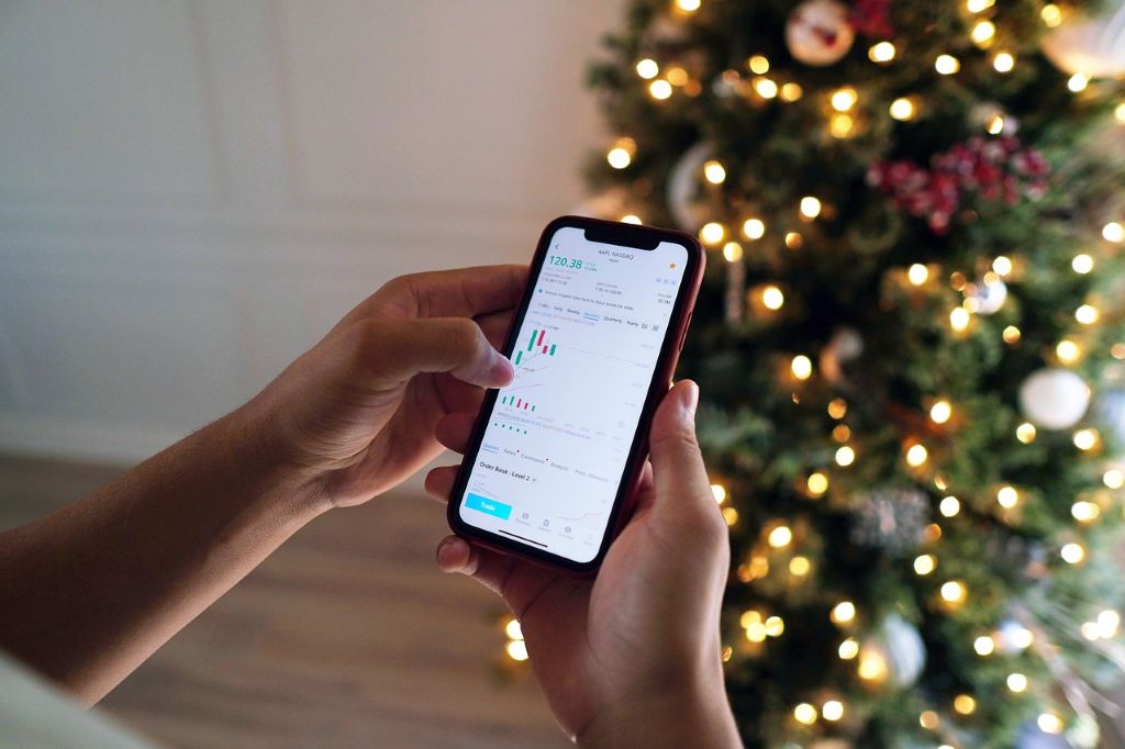 Hände halten ein Smartphone mit Aktien-Charts vor einem geschmückten Weihnachtsbaum – Symbol für Börsengeschäfte während der Feiertage.