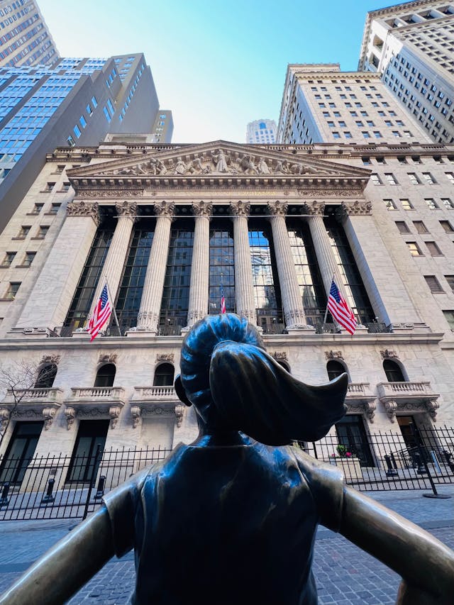 Froschperspektive der Fearless Girl Statue, die furchtlos auf die New York Stock Exchange blickt. Symbolbild für die Entschlossenheit, die finanzielle Freiheit zu erreichen und den amerikanischen Traum – vom Tellerwäscher zum Millionär (Rags to Riches) – wahr werden zu lassen.
