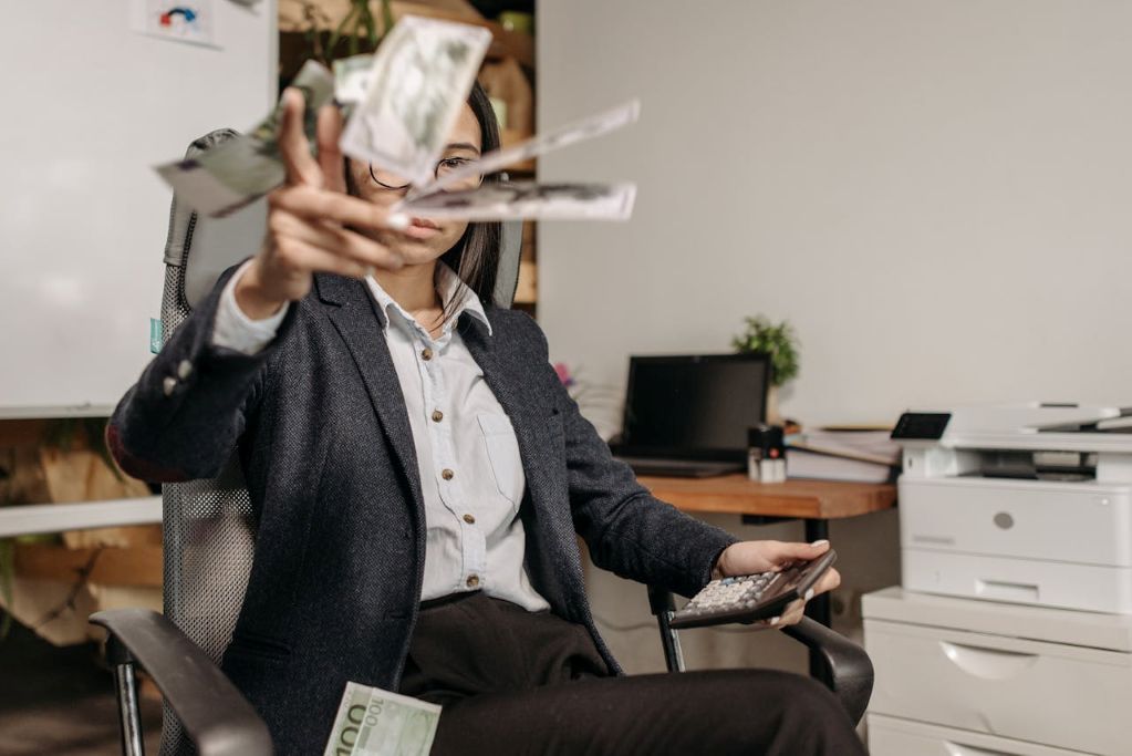 Aufnahme einer Frau in einem Bürostuhl, die mit abgeklärter Mimik Geld in Richtung der Kamera wirft. Ein Symbolbild für echtes Fuck-You-Money und wie man mit System Vermögen erlangt.