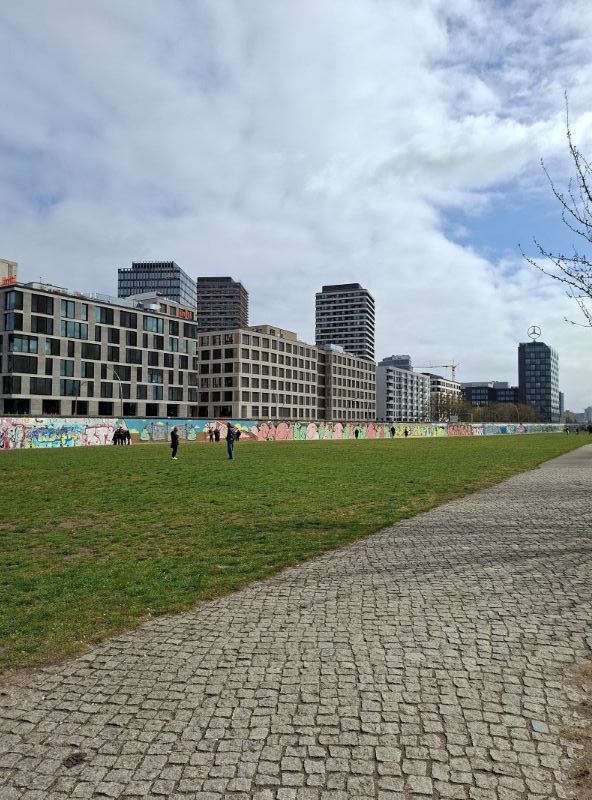 Die bemalte Berliner Mauer an der East Side Gallery mit einer angrenzenden grünen Wiese und modernen Gebäuden im Hintergrund.