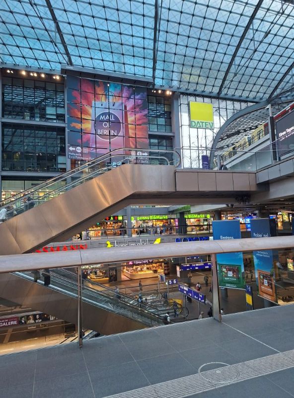 Blick in die moderne, mehrstöckige Glashalle des Berliner Hauptbahnhofs mit Rolltreppen und Geschäften.
