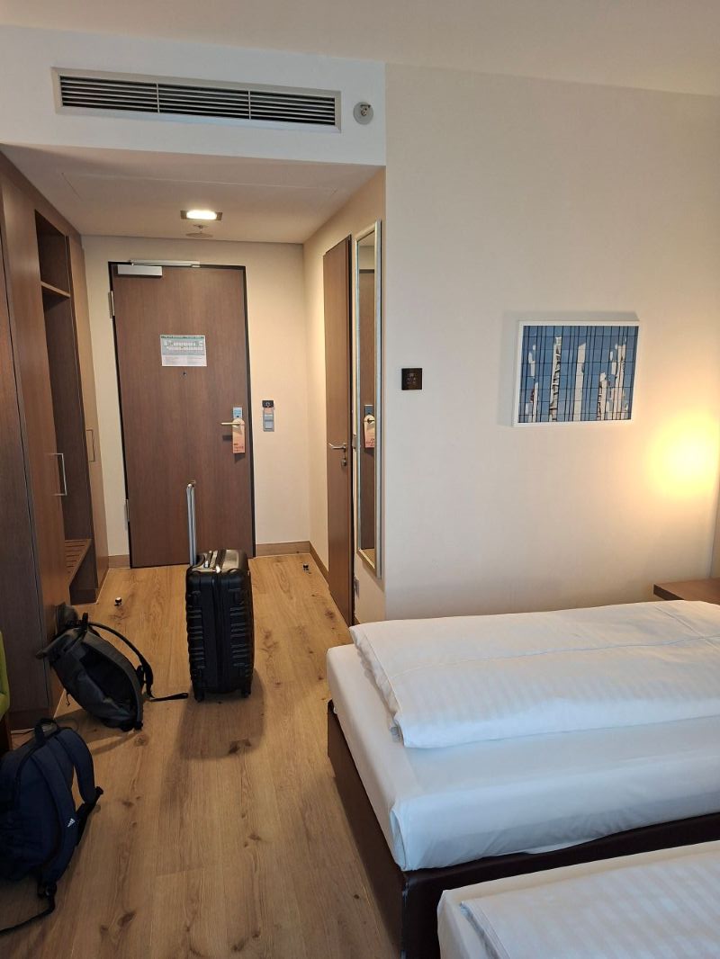 Blick in ein modern und minimalistisch eingerichtetes Hotelzimmer mit einem gemachten Bett und einem abgestellten Koffer im Flurbereich.