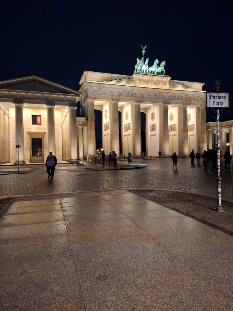 Das beleuchtete Brandenburger Tor am Pariser Platz in Berlin bei Nacht mit einem sichtbaren Straßenschild im Vordergrund.