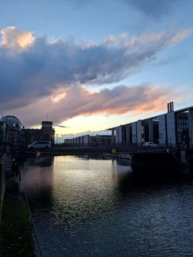 Abendliche Stimmung über der Spree im Berliner Regierungsviertel mit dem Reichstagsgebäude im Hintergrund unter einem farbigen Wolkenhimmel.