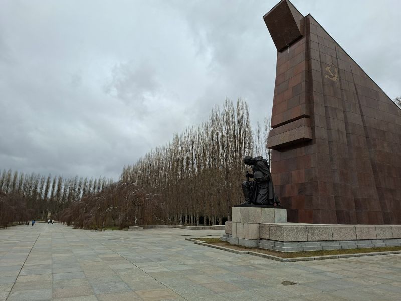 Die monumentale Steinstruktur und eine kniende Soldatenstatue des sowjetischen Ehrenmals im Treptower Park unter wolkenverhangenem Himmel.