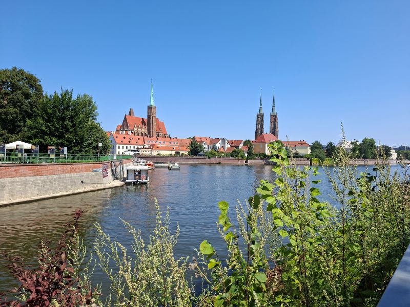 Blick über die Oder auf die historische Altstadt von Breslau mit der St.-Johannes-der-Täufer-Kathedrale unter blauem Sommerhimmel.