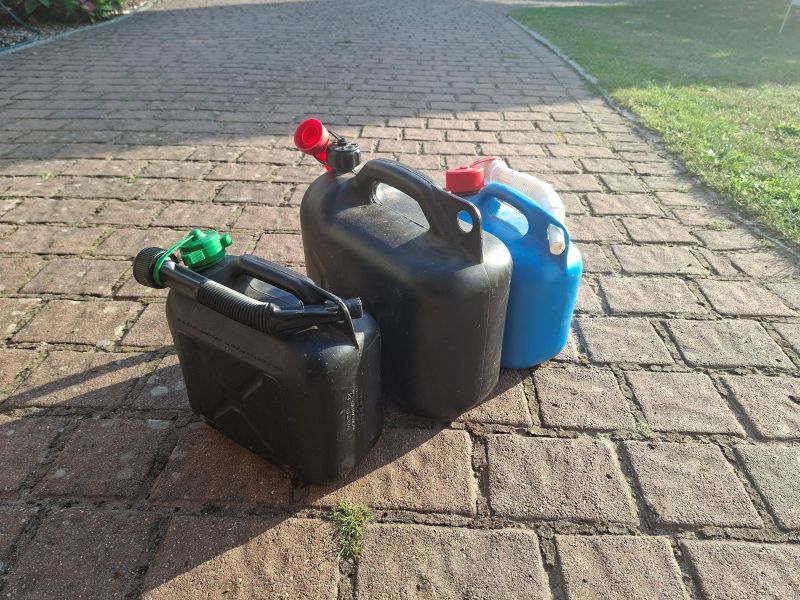 Drei gefüllte Plastik-Benzinkanister aus Schwarz und Blau nebeneinander auf gepflastertem Boden an einem sonnigen Tag.