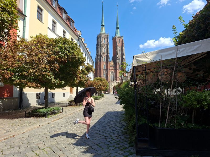 Partnerin des Bloggers posiert mit Regenschirm in einer historischen Pflastergasse vor der Kulisse des Breslauer Doms.