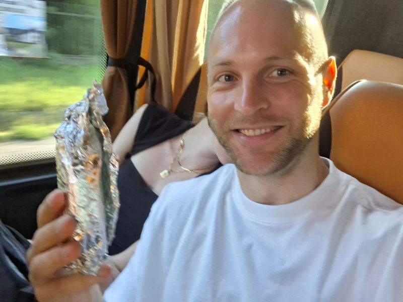 Der Blogger lächelt im Reisebus in die Kamera und hält ein in Alufolie gewickeltes Frühstücksbrötchen hoch.