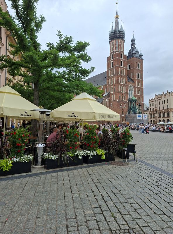 Blick auf die zweitürmige Marienkirche am Hauptmarkt in Krakau mit bestuhlten Außenbereichen im Vordergrund.