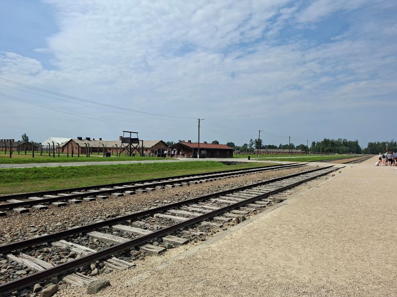 Historische Bahngleise der Selektionsrampe im Vernichtungslager Auschwitz II-Birkenau mit Baracken und Wachturm im Hintergrund.