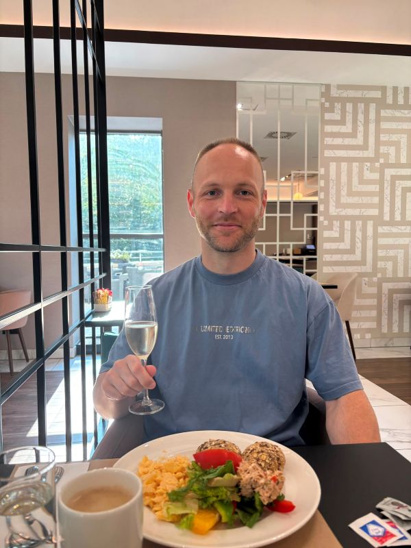 Blog-Autor im blauen T-Shirt sitzt lächelnd mit einem Glas Sekt und einem gefüllten Frühstücksteller am Tisch eines modernen Hotelrestaurants.