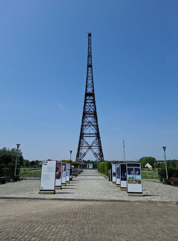 Der historische hölzerne Sendeturm in Gleiwitz mit Informationsstelen im Vordergrund unter blauem Himmel.