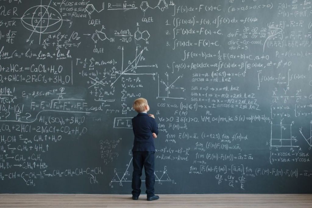 Aufnahme eines kleinen Jungen im Anzug, der in tiefes Nachdenken versunken mathematische Formeln an einer Schultafel studiert. Ein Symbolbild für die Grundlagen des Zinseszinseffekts, Logik und den spielerischen Zugang zur Finanzmathematik.